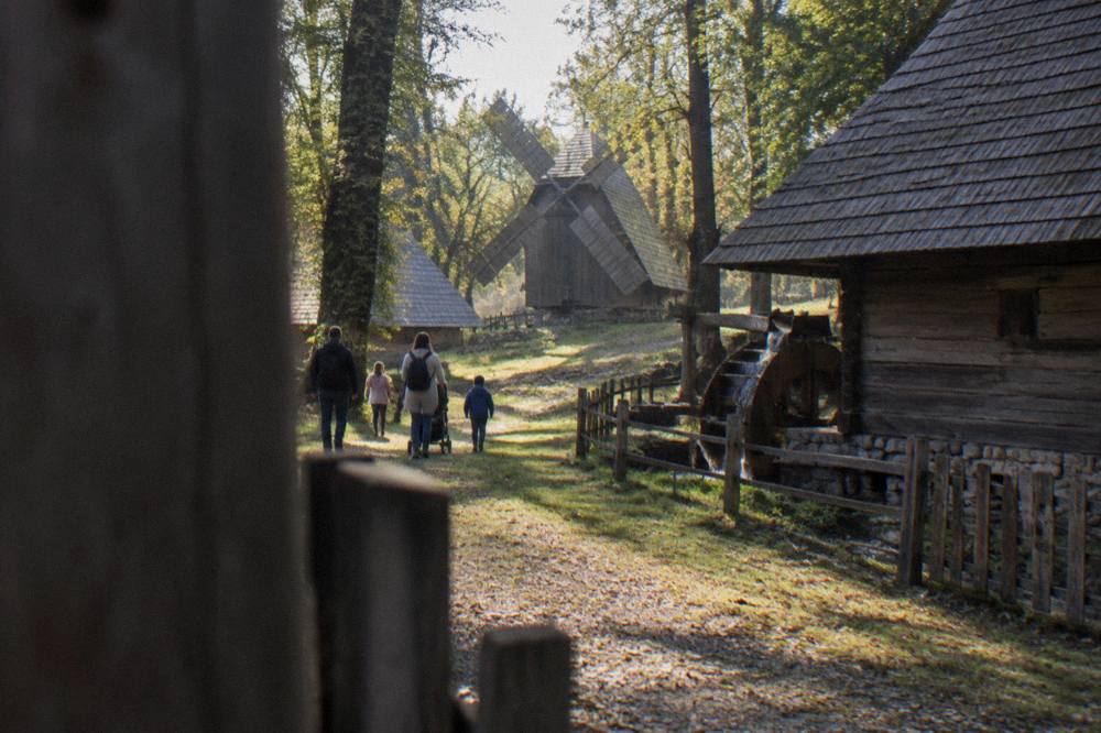 Families walking a forest path beside old wooden farm buildings at an open-air museum