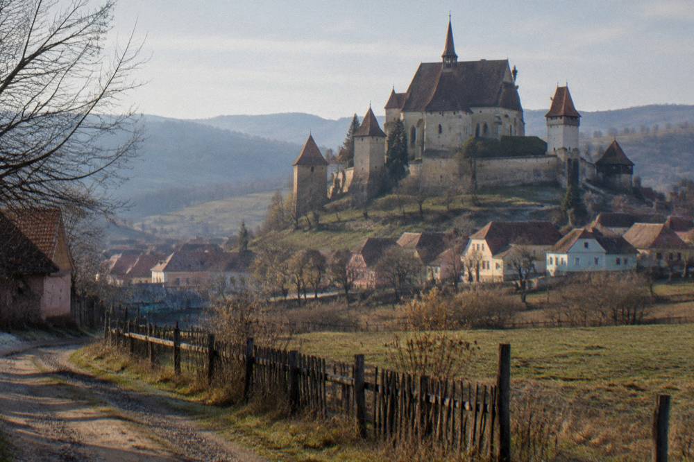 A walled village church with defensive towers seen across farmland in southern Transylvania