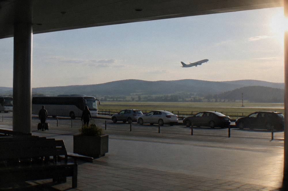 A narrow-body airliner climbing after takeoff above green fields and distant hills