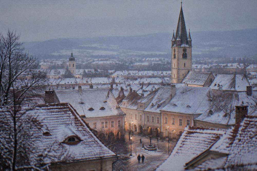 Snow-covered old town rooftops and a church tower seen from a hill at twilight