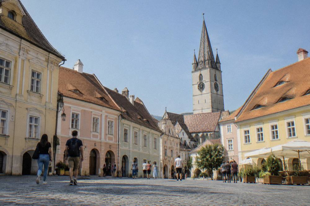 Visitors walking across a small historic square with pastel facades in Sibiu