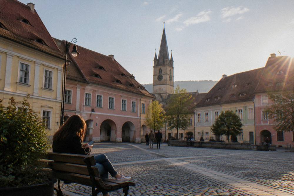 Traveller checking a smartphone on a bench beside pastel-coloured houses in central Sibiu