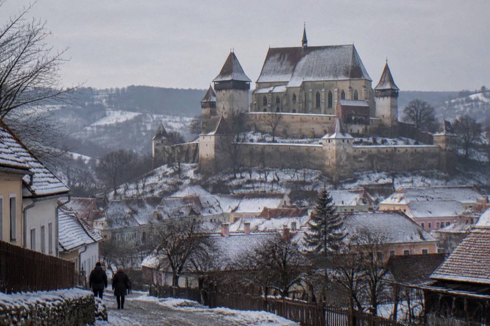 A fortified church with tall defensive walls standing above a snow-covered Transylvanian village