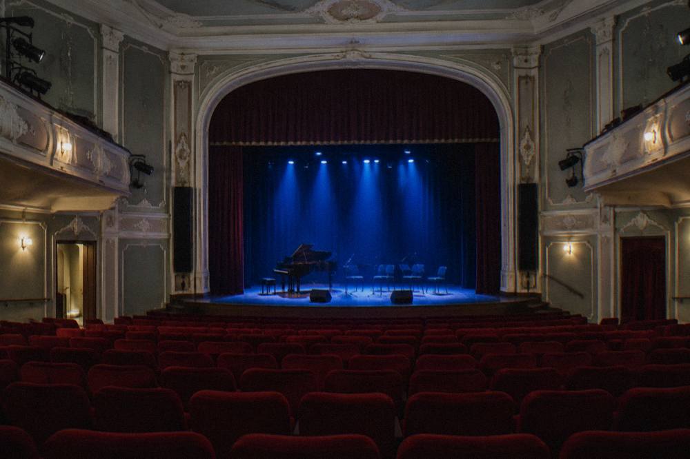 A historic concert hall interior with rows of red seats and an empty stage lit in blue