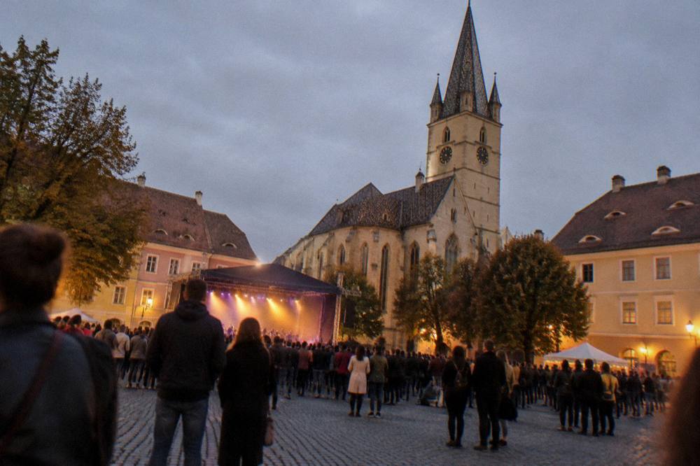A festival crowd in a cobbled square at night with a stage glowing between medieval buildings