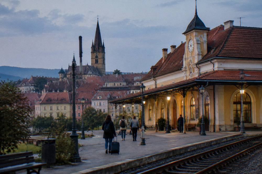 A small train station building with a clock and travellers outside