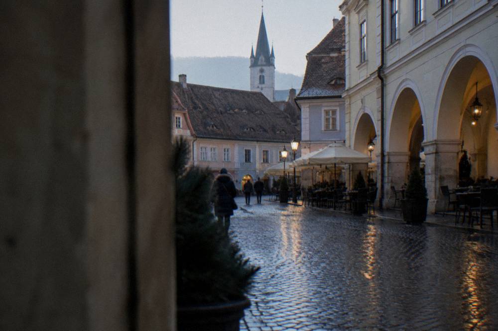 Wet cobblestones and cafe arcades reflecting lamps in a narrow old town square in Sibiu