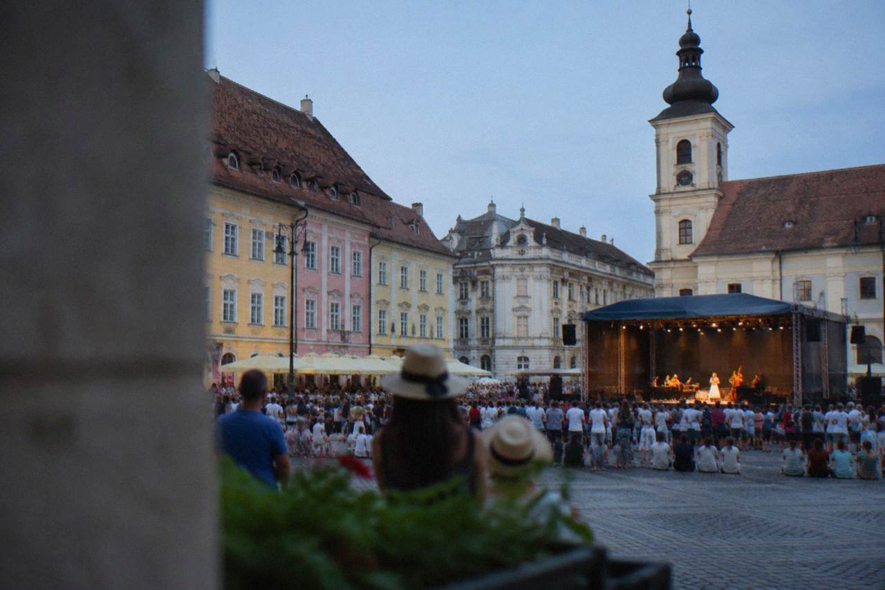 An evening crowd gathered in a large square of a Transylvanian old town watching an open-air stage performance