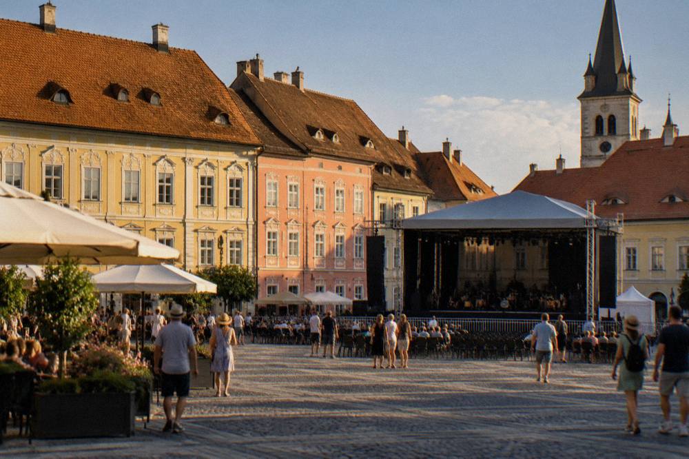 A historic square in Sibiu with colourful facades and a temporary festival stage under a blue June sky
