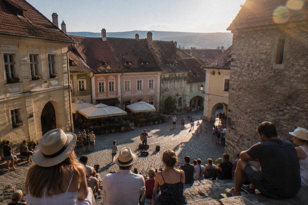 Audience members seated on stone steps beside a medieval tower watching a street performance