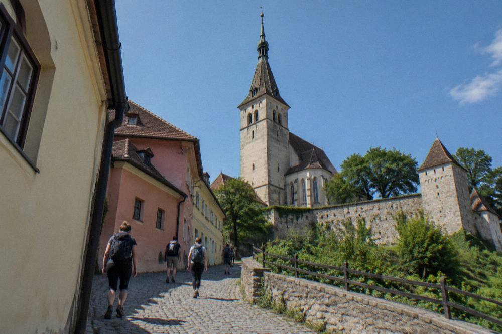 Travellers with backpacks walking up a cobbled lane toward a church tower