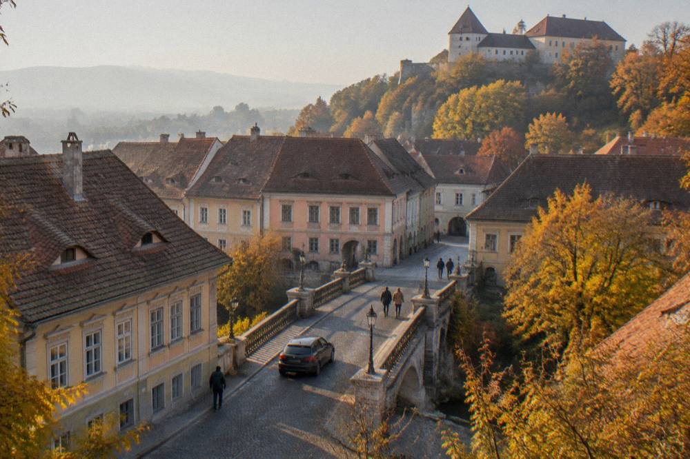 Car crossing a stone bridge toward a hilltop fortress with autumn trees on both sides