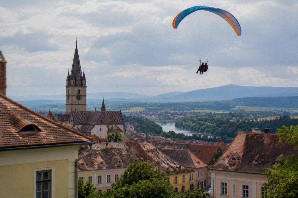 Two riders on a tandem paraglider above green hills and a river valley