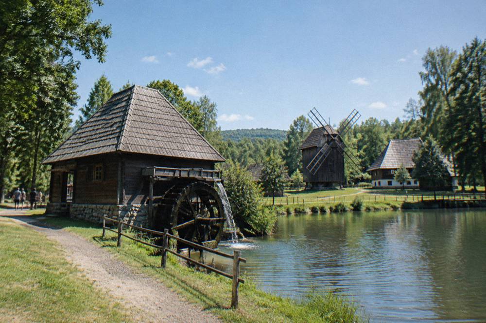 Wooden village houses and a watermill beside a lake in an open-air museum forest