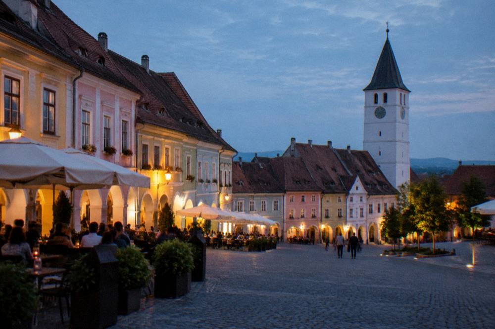 Illuminated Little Square in Sibiu at dusk with people seated at outdoor tables